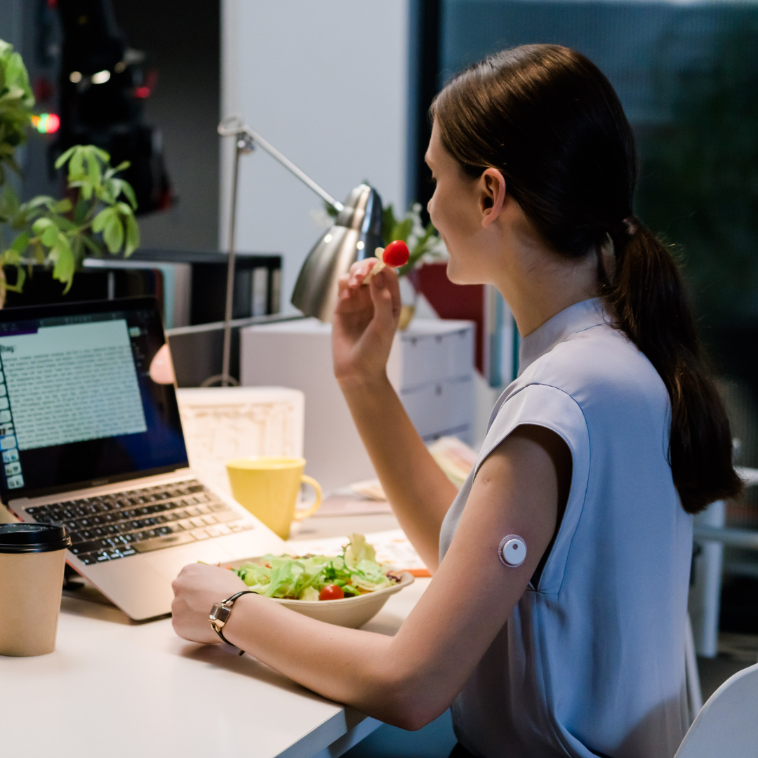 Woman with a CGMS device on her arm eating a salad while working on her laptop at a desk.